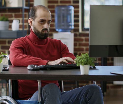 A man in a red sweater sits in a wheelchair working on a computer at a desk, possibly conducting accessibility testing. The background features a brick wall, a water cooler, and shelves with books and plants.