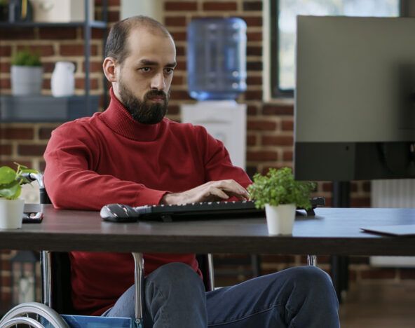 A man in a red sweater sits in a wheelchair working on a computer at a desk, possibly conducting accessibility testing. The background features a brick wall, a water cooler, and shelves with books and plants.