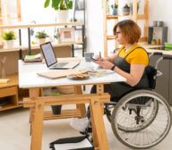 A woman in a wheelchair works on a laptop at a desk in a modern, well-lit home office. She holds a smartphone while surrounded by plants, a notebook, and a plate of cookies. Her focused expression suggests she's engaged in automated accessibility testing.