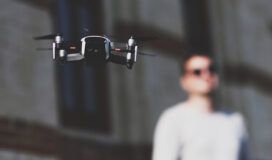 A drone is flying in front of a person wearing sunglasses and standing against a blurred background of a building.