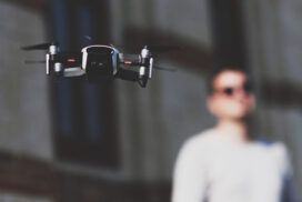 A drone is flying in front of a person wearing sunglasses and standing against a blurred background of a building.