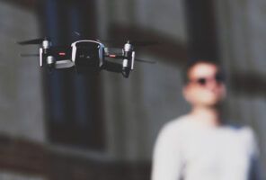 A drone is flying in front of a person wearing sunglasses and standing against a blurred background of a building.