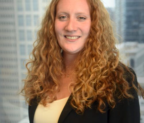 A woman with long curly blonde hair, wearing a yellow top and black blazer, smiles in front of a blurred cityscape background, embodying the principles of test optimization in her confident demeanor.