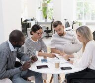 Four people are sitting around a table in a bright office, engaged in a discussion, with papers, notebooks, and electronic devices in front of them.