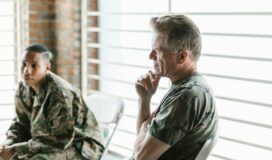 Two people in military uniforms are seated indoors. One is in the foreground, while the other, in the background, is looking away pensively, as if considering how generative AI might influence future strategies.