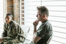 Two people in military uniforms are seated indoors. One is in the foreground, while the other, in the background, is looking away pensively, as if considering how generative AI might influence future strategies.