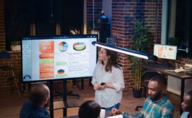 A group of people in a modern office setting are gathered around a presentation screen displaying various charts and data.