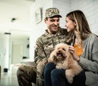 A serviceman in uniform sits on a couch with a woman, both smiling, while she holds a small dog on her lap in a cozy, well-lit room that looks like it was designed by generative AI.