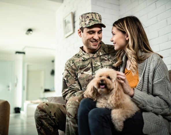 A serviceman in uniform sits on a couch with a woman, both smiling, while she holds a small dog on her lap in a cozy, well-lit room that looks like it was designed by generative AI.