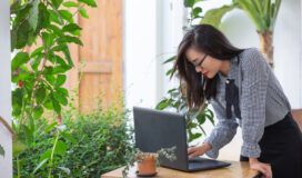 A woman in a checkered shirt works on her laptop at an outdoor table surrounded by plants, skillfully managing Master Data Management tasks.