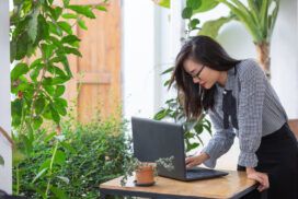 A woman in a checkered shirt works on her laptop at an outdoor table surrounded by plants, skillfully managing Master Data Management tasks.