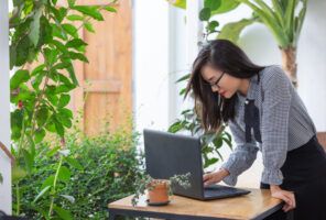 A woman in a checkered shirt works on her laptop at an outdoor table surrounded by plants, skillfully managing Master Data Management tasks.