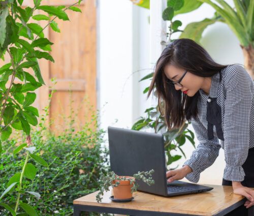 A woman in a checkered shirt works on her laptop at an outdoor table surrounded by plants, skillfully managing Master Data Management tasks.