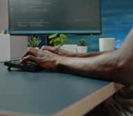 Person typing on a keyboard while working at a desk with a monitor, plants, a cup, and office supplies in the background.