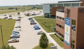 Aerial view of the exterior of a modern, multi-story office building with a sign reading "VGM Group" and a parking lot with multiple cars. Manicured lawns surround the area, and fields are visible in the distance—the hub for innovative machine learning advancements.