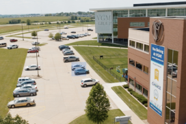 Aerial view of the exterior of a modern, multi-story office building with a sign reading "VGM Group" and a parking lot with multiple cars. Manicured lawns surround the area, and fields are visible in the distance—the hub for innovative machine learning advancements.