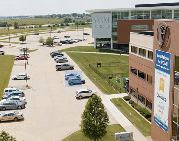 Aerial view of the exterior of a modern, multi-story office building with a sign reading "VGM Group" and a parking lot with multiple cars. Manicured lawns surround the area, and fields are visible in the distance—the hub for innovative machine learning advancements.