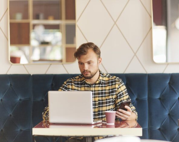 A man with a beard is sitting in a cafe, focused on his laptop while holding a smartphone in his other hand. There is a paper coffee cup on the table in front of him.