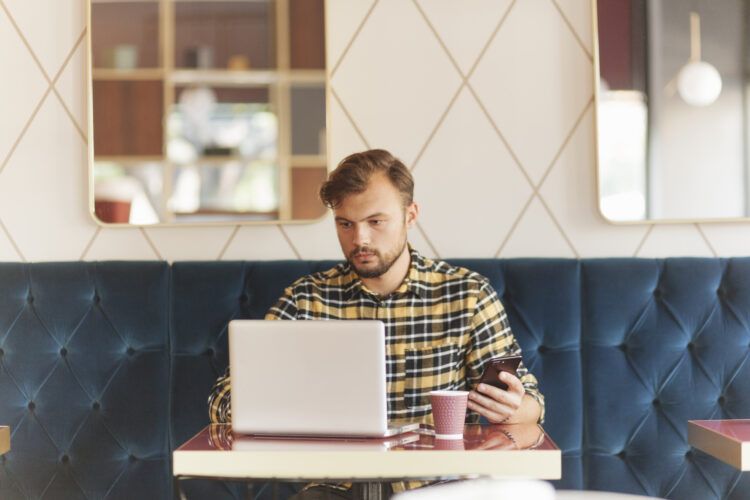 A man with a beard is sitting in a cafe, focused on his laptop while holding a smartphone in his other hand. There is a paper coffee cup on the table in front of him.