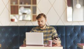 A man with a beard is sitting in a cafe, focused on his laptop while holding a smartphone in his other hand. There is a paper coffee cup on the table in front of him.