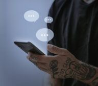 A person with tattooed arms holding a smartphone, with three chat bubbles above the phone.