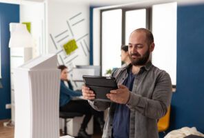 A man with a tablet stands in a modern office, with two coworkers in the background working at desks.