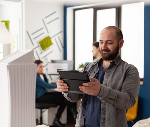 A man with a tablet stands in a modern office, with two coworkers in the background working at desks.