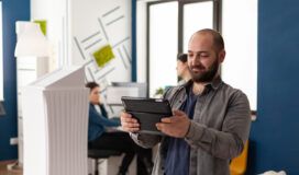 A man with a tablet stands in a modern office, with two coworkers in the background working at desks.