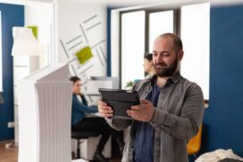 A man with a tablet stands in a modern office, with two coworkers in the background working at desks.