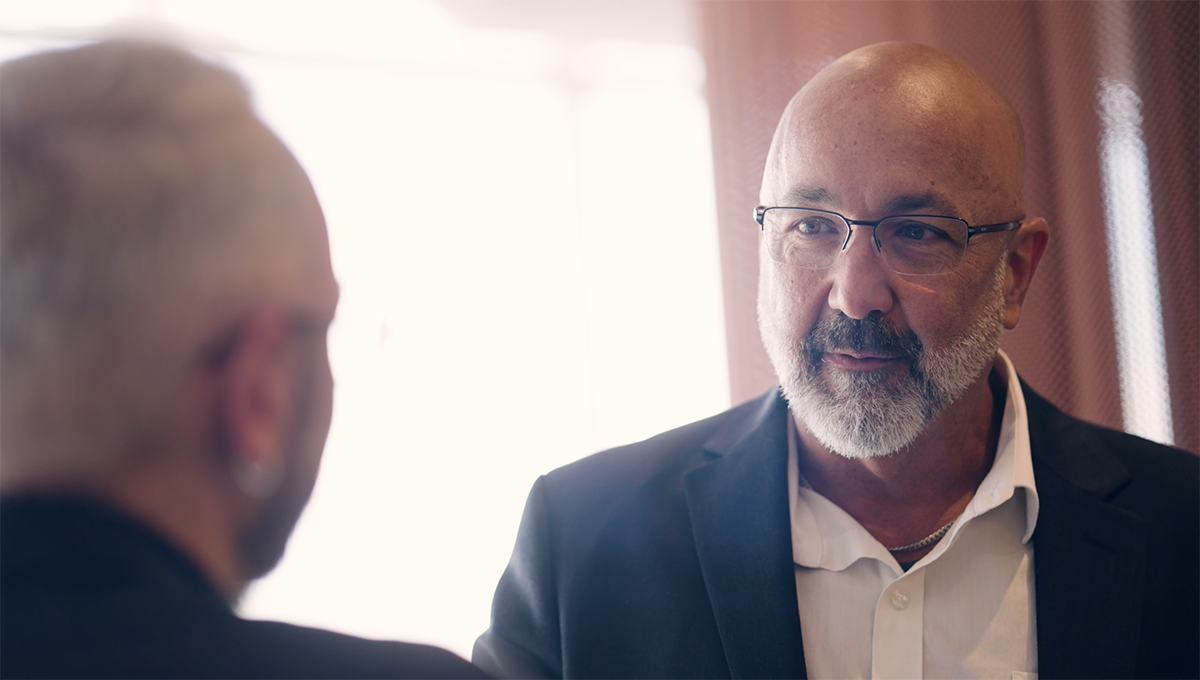 Two men in suits are engaged in a conversation. One, facing the camera with glasses and a white beard, appears thoughtful. The other, blurred from the back, seems to be discussing machine learning innovations.