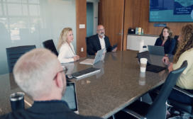 Five people are seated around a conference table with laptops, discussing a project in a modern office room. A digital screen on the wall displays information about machine learning advancements.