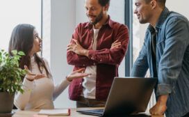 Three colleagues engaged in conversation about AI for project management in an office. A woman seated at a desk gestures as two men stand nearby, one with arms crossed and the other leaning on the desk. A laptop and plant are present.