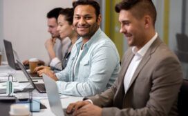 Four people sitting in a row at a conference table working on laptops, possibly using AI for project management. One person is smiling at the camera.