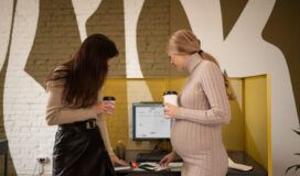 Two women, one pregnant, stand by an office desk with coffee cups, reviewing documents on a desk; a computer monitor shows an AI for project management calendar in the background.