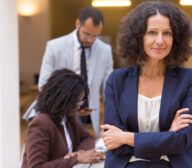 A confident woman in a business suit stands with her arms crossed, overseeing as her two colleagues work on integrating AI for project management in the background.