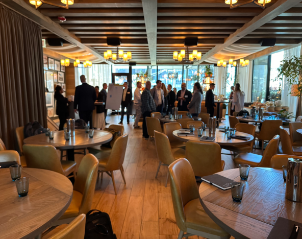 A group of people gathers near a presentation board in a warmly lit restaurant with round wooden tables and yellow chairs.