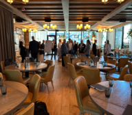 A group of people in business attire stand and converse in a restaurant with round tables, wooden floors, and overhead lighting.