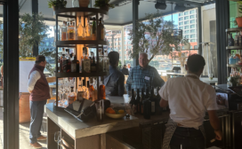 People are gathered around a restaurant bar, with bottles and glasses displayed. Large windows provide a view of the city buildings outside.