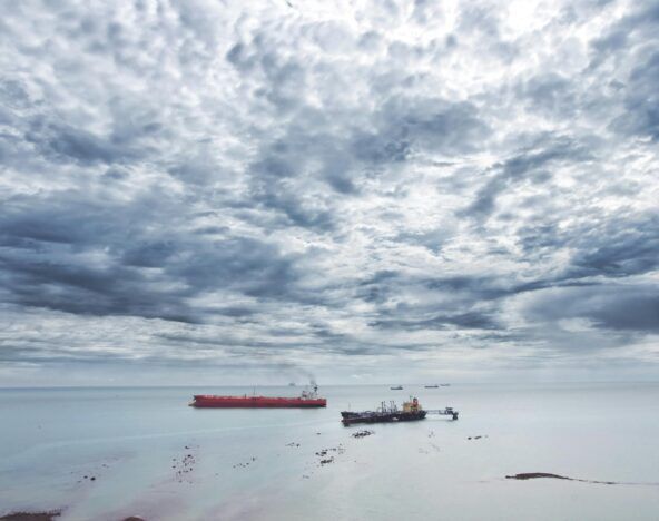 A cloudy sky over a calm sea with a red cargo ship and smaller vessels in the distance.