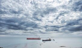 A cloudy sky over a calm sea with a red cargo ship and smaller vessels in the distance.