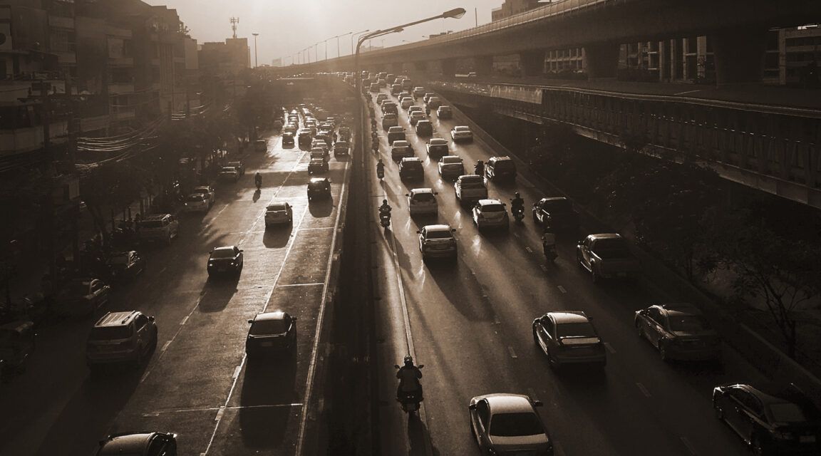 A city highway in sepia tone with heavy traffic under a setting sun; elevated train tracks run parallel to the road on the right.