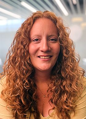 A person with long, curly red hair smiles at the camera in a softly lit indoor setting.