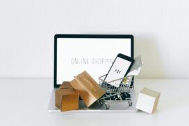 A small shopping cart with a smartphone and credit card sits on a laptop, surrounded by small packages. The laptop screen displays the text "ONLINE SHOPPING.