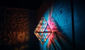 Geometric metal sculpture on a textured wall casts colorful shadows with blue and orange lighting.