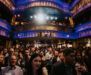 A large crowd of people gathered inside an ornate theater with blue and gold balconies.