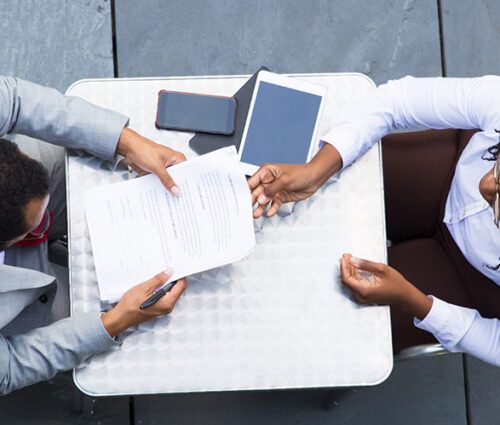 Two people seated at a table with documents, a tablet, and smartphones on it. They appear to be discussing or reviewing the paperwork.