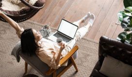 Woman relaxed in chair with a laptop on her lap, holding a mug. One arm is raised. Room features wooden flooring, a plant, and cozy furniture.