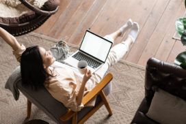 Woman relaxed in chair with a laptop on her lap, holding a mug. One arm is raised. Room features wooden flooring, a plant, and cozy furniture.