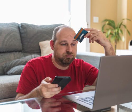A man in a red shirt sits in front of a laptop, holding a credit card to his forehead and a phone in his other hand, appearing frustrated as he struggles with capacity planning.