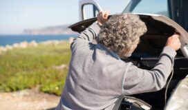 Older man with gray curly hair inspects the engine under the hood of a vehicle, exploring technology integration, parked outdoors near a grassy area with the sea in the background.